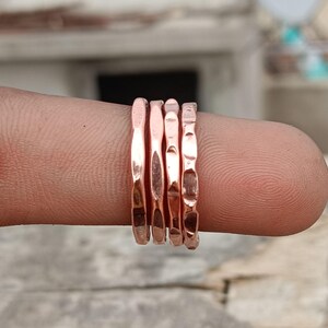 May include: A set of four copper-coloured rings with a hammered, textured finish. Each ring features a distinct, irregular surface pattern with reflective metallic highlights, showcasing a polished appearance and a slim, stackable band design.