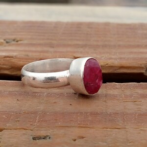 Pu&ograve; includere: Un anello d'argento con una grande gemma ovale di colore rosso intenso. La fascia dell'anello &egrave; liscia e semplice, e la gemma &egrave; incastonata in una lunetta. L'anello &egrave; esposto su una superficie di legno.