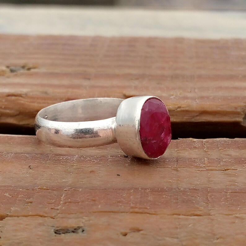 Pu&ograve; includere: Un anello d'argento con una grande gemma ovale di colore rosso intenso. La fascia dell'anello &egrave; liscia e semplice, e la gemma &egrave; incastonata in una lunetta. L'anello &egrave; esposto su una superficie di legno.