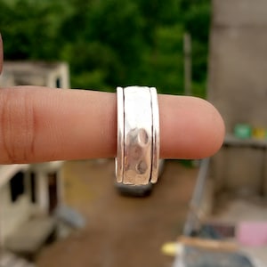 May include: A silver ring with a hammered texture. The ring has a wide band with raised edges. The ring is held on a finger, with a blurred green background. The ring's design is approximately 1 cm wide.