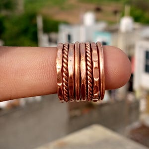 May include: A collection of copper-colored rings with various textures, including twisted rope and hammered designs. The rings are stacked on a finger, showcasing their different styles and finishes. The rings are a warm, metallic color.