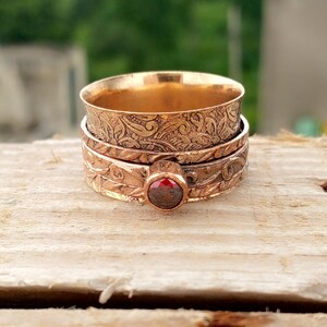 May include: A copper-colored spinner ring with intricate floral patterns. The ring features a central band with a small, round, red gemstone. The design includes detailed engravings, creating a vintage or antique aesthetic.