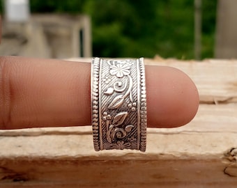 Puede incluir: Un anillo de plata con un intrincado diseño floral. El anillo presenta tallas detalladas de flores, hojas y vides, con un borde texturizado. El anillo se muestra en un dedo sobre un fondo verde borroso.