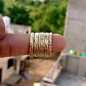 Puede incluir: Una colección de seis anillos dorados, cada uno con un diseño en relieve único. Los anillos están apilados en un dedo, mostrando los intrincados patrones y texturas. El fondo está borroso, con follaje verde y un edificio visible.
