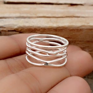 May include: A silver-colored ring with a layered design. The ring features multiple bands that intersect and overlap, creating a textured appearance. The ring is shown on a hand, with a wooden surface in the background.