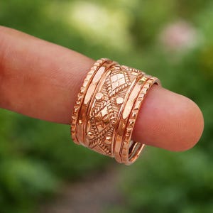May include: A collection of rose gold rings, each with detailed geometric patterns and textures. The rings are displayed on a finger, highlighting the intricate design and warm metallic hue. The rings are a jewelry item.