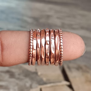May include: A collection of copper-colored rings stacked on a finger. The rings have varying textures, including smooth, ridged, and hammered designs. The rings are a warm, reddish-brown hue, and the image is well-lit.