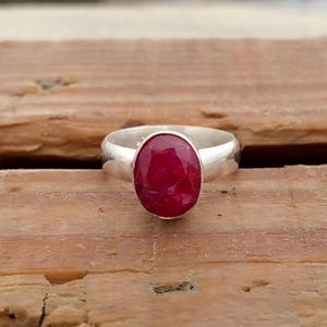 Pu&ograve; includere: Un anello d'argento con una grande gemma di rubino ovale. Il rubino &egrave; di un rosso intenso ed &egrave; incastonato in una fascia d'argento lucida. L'anello &egrave; esposto su una superficie di legno. Il design &egrave; semplice ed elegante.