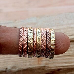 Op de afbeelding: Een set van acht dunne metalen ringen op een rij, met diverse texturen en patronen zoals stippen, zigzaglijnen, bloemmotieven en bladontwerpen. De ringen hebben een mix van koper-, brons- en messingkleurige metalen afwerkingen.