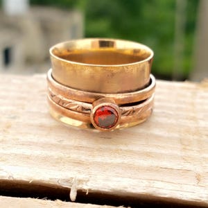 May include: A gold-toned spinner ring with a red gemstone. The ring features a wide band and several smaller bands, one with an etched design. The gemstone is oval-shaped and set in a bezel. The ring is displayed on a wooden surface.