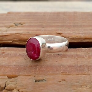 Pu&ograve; includere: Un anello d'argento con una grande pietra preziosa rosso scuro di forma ovale. L'anello ha una fascia semplice e un'incastonatura a castone che tiene saldamente la pietra. L'anello &egrave; esposto su una superficie di legno.