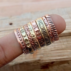 Peut inclure: Une collection de bagues en cuivre et dorées avec divers motifs en relief, notamment des cœurs, des feuilles et des motifs géométriques. Les bagues sont empilées sur un doigt, mettant en valeur les différentes textures et couleurs.