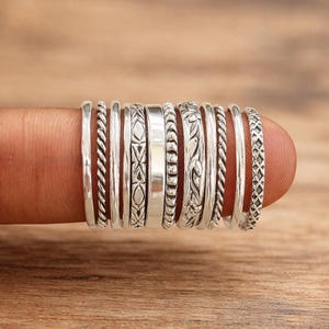 Puede incluir: Una colección de anillos de plata con varios diseños, incluyendo bandas trenzadas, estampadas y lisas. Los anillos se muestran en un dedo, mostrando los diferentes estilos y texturas. Los anillos son de plata.