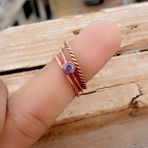 May include: A set of three copper rings. One ring has a twisted design, another has a small, round, purple gemstone, and the third is a simple band. The rings are stacked on a finger.
