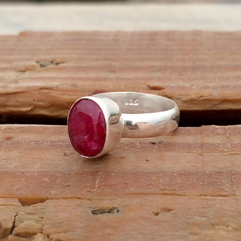 Pu&ograve; includere: Un anello d'argento con una grande pietra preziosa rosso scuro di forma ovale. La fascia dell'anello &egrave; liscia e lucida e la gemma &egrave; incastonata in un'incastonatura a castone. L'anello &egrave; esposto su una superficie di legno.