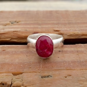 May include: A silver ring featuring a large, oval-cut ruby gemstone. The ruby is a deep, vibrant red color and is set in a simple, polished silver band. The ring is displayed on a wooden surface.