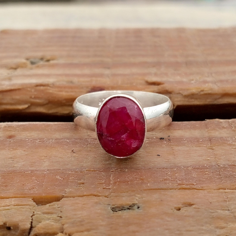 Pu&ograve; includere: Un anello d'argento con una grande gemma di rubino ovale. Il rubino &egrave; di un rosso intenso ed &egrave; incastonato in una fascia d'argento lucida. L'anello &egrave; esposto su una superficie di legno. Il design &egrave; semplice ed elegante.