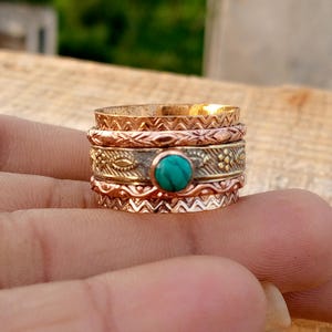 May include: A collection of mixed-metal rings with detailed patterns. The rings, stacked on a finger, include a central ring with a turquoise stone. The rings are made of copper and gold-toned metal, with decorative engravings.