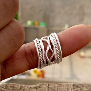 Op de afbeelding: Een zilveren ring met meerdere banden en een gevlochten ontwerp. De ring, gedragen op een vinger, heeft een gedetailleerd, getextureerd uiterlijk. Het ontwerp bevat gladde en gedraaide elementen, wat een unieke uitstraling creëert.
