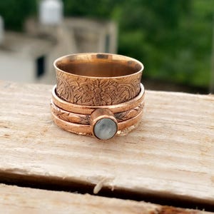 May include: A copper-colored spinner ring with floral and geometric patterns. The ring features a light blue gemstone and is made of multiple bands that rotate independently. The ring is displayed on a wooden surface.