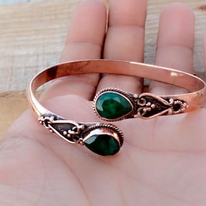Op de afbeelding: Koperen manchetarmband met twee druppelvormige, levendige groene edelstenen. De armband heeft een open ontwerp met sierlijke details rond de edelstenen. Het koper heeft een warme, roodbruine tint, wat contrasteert met de groene stenen.