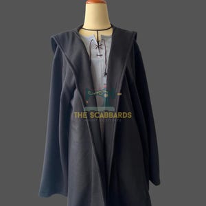 May include: A black hooded cloak over a light gray tunic with a black choker. The tunic has a lace-up neckline. The text "THE SCABBARDS ANIME COSTUMES" is visible on the front of the tunic.