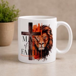 May include: A white ceramic mug featuring a graphic design that splits a dark cross and a detailed lion face. The text "MAN of FAITH" is printed in black lettering on the left side of the cross, with orange watercolor accents surrounding the imagery.