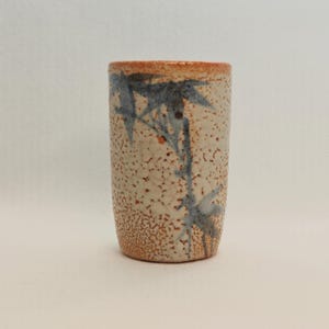 May include: A cylindrical ceramic cup with a speckled beige exterior and a brown rim. The cup features a blue bamboo design. The cup is approximately 4 inches tall.