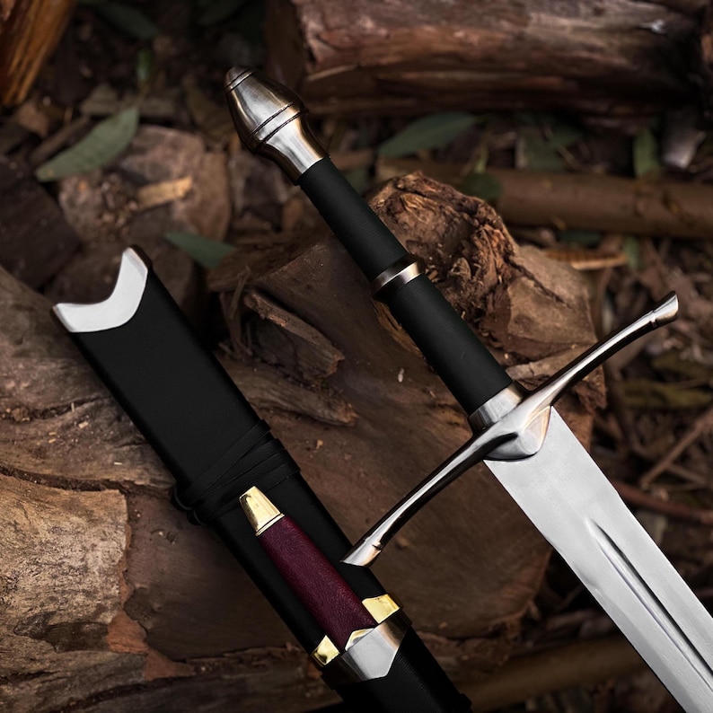 Op de afbeelding: Een middeleeuws zwaard met een zilveren kling en pareerstang, een zwarte handgreep en een zilveren pommel. Het zwaard wordt getoond met zijn zwarte schede, die een bordeauxrode en gouden accent heeft. Het zwaard rust op een bed van hout.