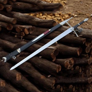 Op de afbeelding: Een lang, zilveren zwaard met een zwarte handgreep en een zilveren pareerstang, naast een zwarte schede met een klein mes. Het zwaard en de schede liggen op een stapel bruine boomstammen. Het mes heeft een rood-bruine handgreep.