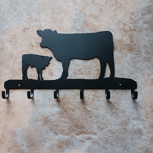 May include: Black metal wall hook featuring a cow and calf silhouette design. The hook has five curved hooks for hanging items. A decorative and functional piece for a kitchen or entryway.
