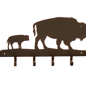 May include: A dark brown metal wall-mounted coat rack featuring a silhouette of a bison and calf. The rack has five hooks for hanging items. The bison and calf are walking on a horizontal bar.