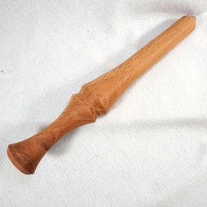 May include: A handcrafted wooden wand with a light brown finish. The wand features a tapered handle and a decorative, geometric design near the center. The wand is approximately 12 inches long.