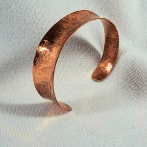 May include: A hammered copper cuff bracelet with a wide, curved band. The bracelet is made of a single piece of copper that has been hammered to create a textured surface.
