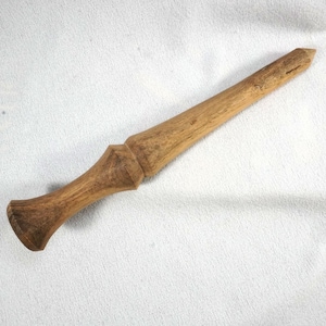 May include: A handcrafted wooden wand with a light brown finish. The wand features a flared handle, a decorative midsection, and a tapered tip. The wand is displayed on a white textured surface.