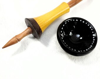 May include: A wooden spindle with a yellow body and a dark brown accent, paired with a small, black bowl with white dotted trim. The spindle has a pointed end and a rounded top. The bowl is round and glossy.
