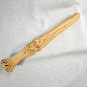 May include: A light-colored wooden wand with a carved handle and a tapered body. The handle features a diamond-shaped design. The wand is approximately 12 inches long and has a smooth, polished finish. The wand is made of a light-colored wood with visible grain.