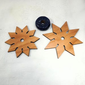 Puede incluir: Dos posavasos de madera en forma de estrella con un agujero central y puntas recortadas. También hay un anillo de cerámica azul oscuro. Los posavasos son de madera clara y el anillo de cerámica tiene una superficie texturizada.