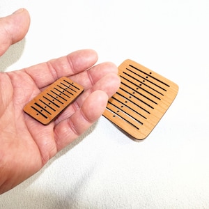 May include: Two small, light brown wooden objects with parallel grooves and small holes. One is rectangular, the other square. The objects are held in a hand against a white background.