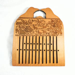 Op de afbeelding: Een lichtgekleurde houten weefkam met een handvat en een decoratief gesneden ontwerp met dieren en geometrische patronen. De kam heeft verticale sleuven en kleine gaten.