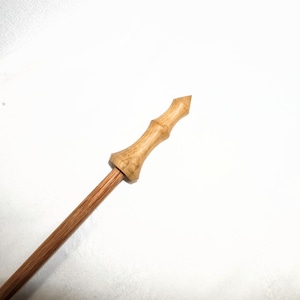 May include: A wooden wand with a light brown handle and a darker brown shaft. The handle has a carved design and a pointed tip. The wand is angled diagonally across the frame against a white background.