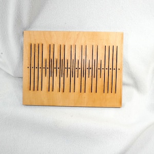 May include: A rectangular wooden plaque with a series of vertical black lines and small dots. The lines vary in height, creating a visual pattern. The wood has a light, natural color.