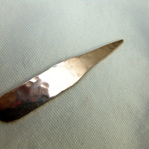 Könnte beinhalten: Ein gehämmerter silberner Brieföffner mit einer spitzen Spitze. Der Brieföffner liegt auf einem weißen Stoffhintergrund.