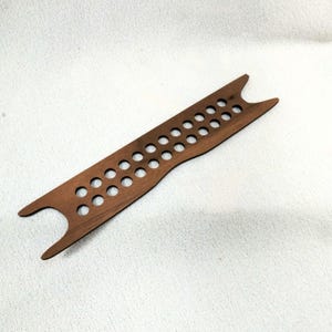 May include: A wooden weaving shuttle with a series of evenly spaced holes and a forked end. The shuttle is a warm brown color and appears to be made of a single piece of wood. It is designed for use in textile crafts.