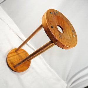 May include: A handmade wooden stand with a circular base and top, linked by two thin wooden supports. The top has a central hole and is decorated with inlaid elements. The wood has a warm, natural finish.