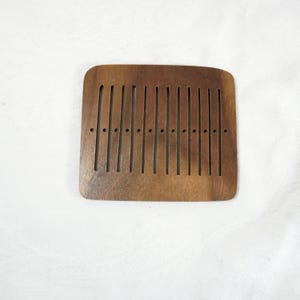 May include: A square wooden comb with a dark brown finish. The comb has a series of vertical slots and small holes along the top edge. The comb is designed for hair care and styling.