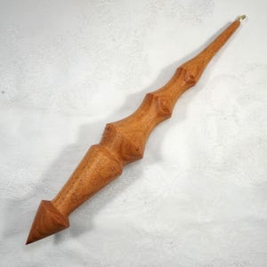May include: A handcrafted wooden wand with a pointed tip and a decorative, textured design. The wand is a light brown colour and has a small gold-coloured loop at the top. The wand is approximately 30 cm long.