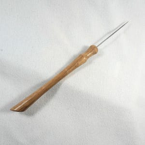 Könnte beinhalten: Ein Holzwerkzeug mit einer langen, dünnen Metallnadel, die an einem Ende befestigt ist. Das Werkzeug hat einen glatten, abgerundeten Holzgriff.