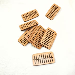 Könnte beinhalten: Eine Sammlung kleiner, rechteckiger Holzstücke mit parallelen Schlitzen und gleichmäßig verteilten Löchern. Die Stücke sind hellbraun und scheinen lasergeschnitten zu sein. Die Objekte sind auf einer weißen Oberfläche verstreut.