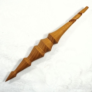 May include: A handcrafted wooden spindle with a pointed end and a decorative, geometric design. The spindle is light brown and features a spiraled top section. The overall length is approximately 8 inches.